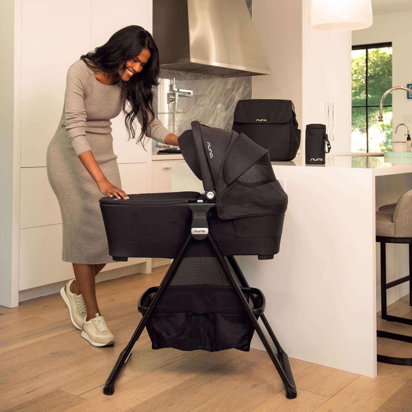 Nuna Diaper Bag - Caviar sits on counter while mom looks at baby in Nuna bassinet