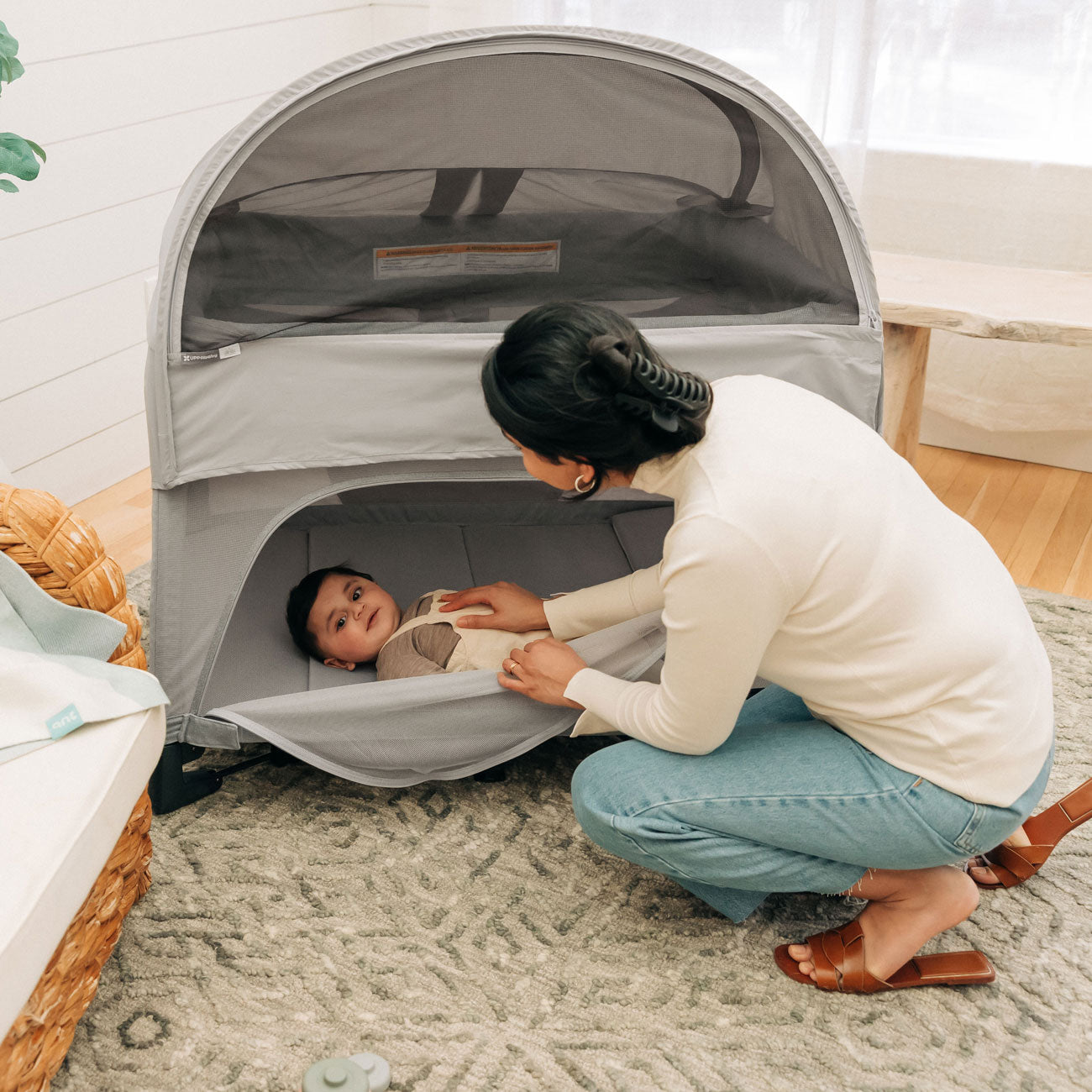 Mom checks on baby lying in REMI playard with attached UPPAbaby REMI Playard Canopy