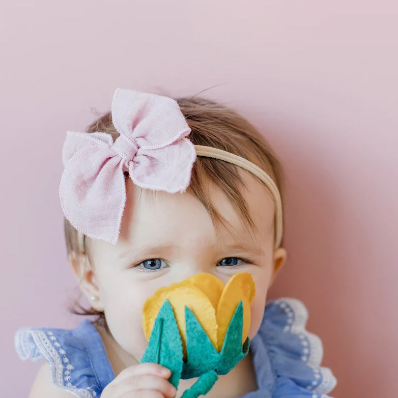 Baby girl holding flower wears Modern Piggy Gauze Petite Party Bow - Nylon Headband - Blush