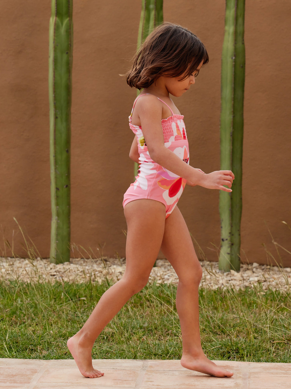 Girl walking wears Girl sits by pool wearing Tea Collection Smocked One-Piece Swimsuit - Floral Burst 